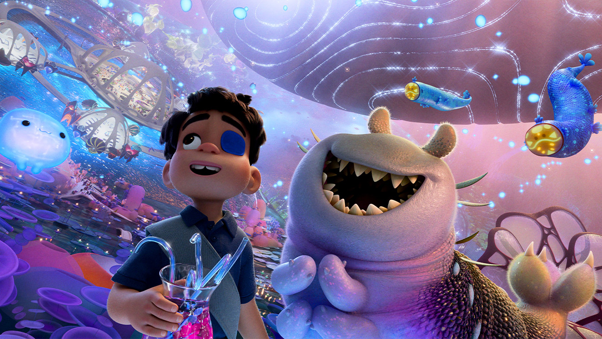 In a still from Disney and Pixar’s Elio, Elio (voiced by Yonas Kibreab), left, and Glordon (voiced by Remy Edgerly), right, are standing in the middle of the Communiverse and taking in the incredible view around them. Glordon is an adorable alien of the Hylurgian species, which looks like a worm with giant teeth, no discernable eyes, and several arms. He has a huge smile on his face. Elio looks happy too; he has an eyepatch over one eye and is holding a glass of Glorp, the drink of choice in the Communiverse. Ooooo the supercomputer can be seen to Elio’s left.