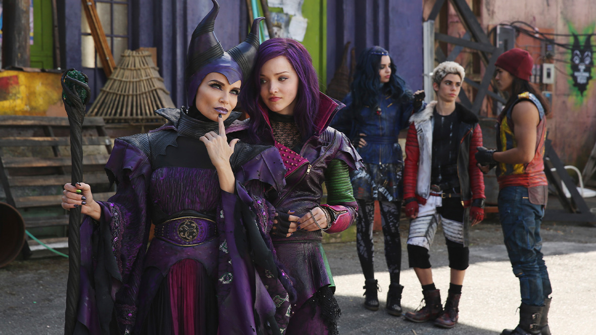 In a still from Descendants (2015), Mal (Dove Cameron) is standing with her mother Maleficent (Kristin Chenoweth) on the left, while Sofia Carson as Evie, Booboo Stewart as Jay, and Cameron Boyce as Carlos stand in the background off to the right, talking amongst themselves.