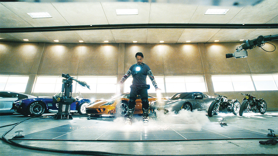Image of Tony Stark testing out Iron Man technology in Iron Man (2008). He is standing in front of a row of sport cars and motorcycles. He is flanked on either side by two robotic arms, DUM-E (left) and U (right), which are recording his test of his Iron Man technology.