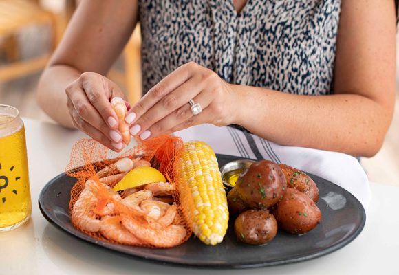 A woman wearing a diamond ring cracks open a fresh shrimp over a black plate housing an ear of corn, seasoned potatoes, and a net with more shrimp. Next to the plate is a glass filled with cold beer sporting a “P” logo. 