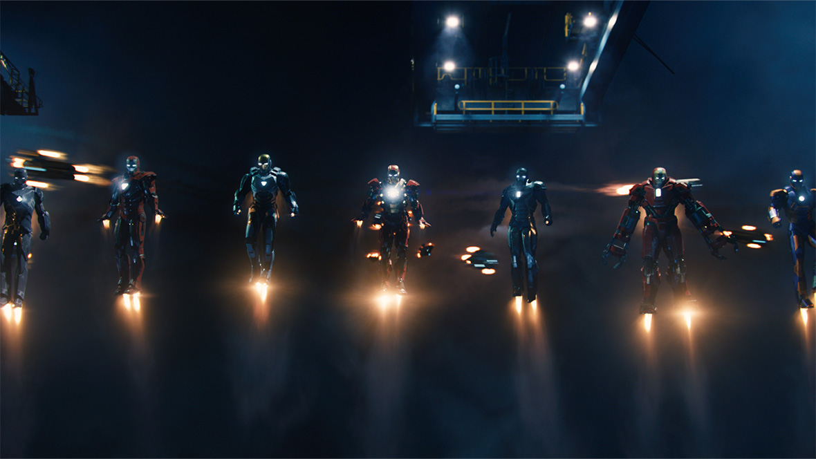 Seven Iron Man suits, part of the Iron Legion, hovering above the ground in front of a dark blue background.
