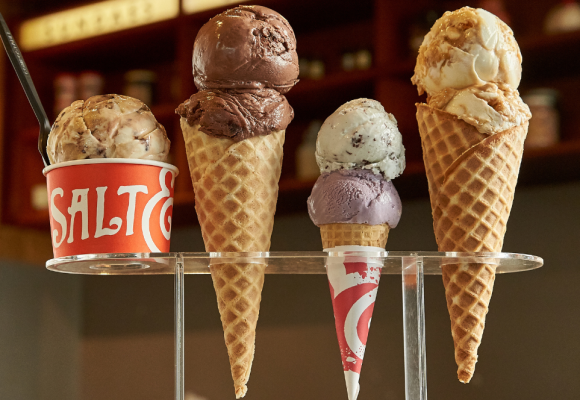 A red cup of ice cream and three ice cream cones (all varying in size) sit housed in an ice cream cone holder. The red cup of ice cream adorns the Salt & Straw logo and contains a brown & white colored ice cream (with chocolate fudge mixed in). The ice cream cones all contain two scoops of ice cream that range in flavor. 