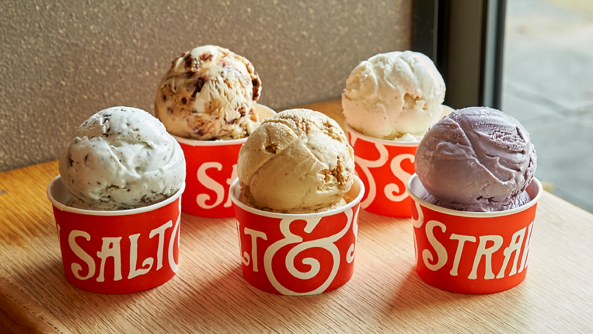 Gold Member Offer: 10% Off at Salt & Straw in Downtown Disney, CA & Disney Springs, FL Five various red cups of frozen ice cream with the Salt & Straw logo rest on a light wooden table near a window. The ice cream housed in the cups range in terms of color & flavor. Two are brown, two are white, and one is a light purple. Two of the ice cream cups have chocolate toppings mixed into them.