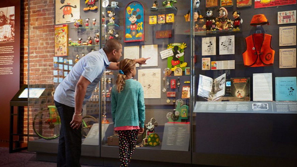 Gold Member Offer: Save $3 on General Admission at The Walt Disney Family Museum, San Francisco A father and daughter stand admiring a display at The Walt Disney Family Museum. The wall is filled with Mickey Mouse memorabilia including watches, clocks, figurines, dolls, books, illustrations, and more. The dad is pointing to a display from behind the protective glass.