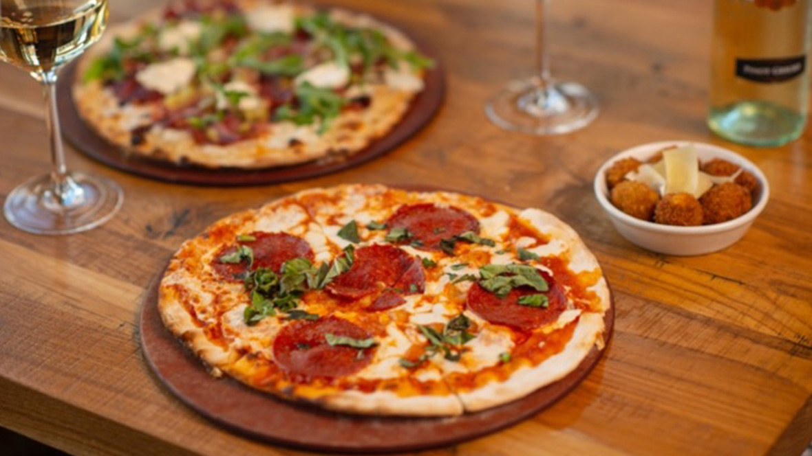 Gold Member Offer: 10% Off at Terralina Crafted Italian, Disney Springs A fresh thin-crust pepperoni pizza sits atop a round serving tray. The cheese is perfectly melted, and the pizza is topped with a green garnish. Behind this pizza sits a different type of pizza pie just out of focus. On the left and right are two glasses filled with white wine.