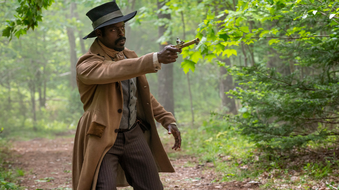 Sterling K. Brown as Medwin Harris, standing in the woods and aiming a small gun at something off-screen to the right.