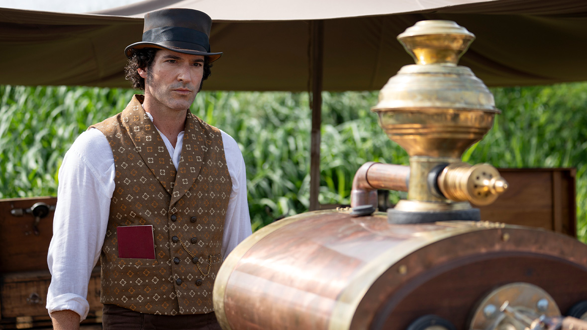 Tom Ellis, as Christopher ‘Titch’ Wilde, stands beside an umbrella, staring a machine that is in the foreground and partially out of focus.
