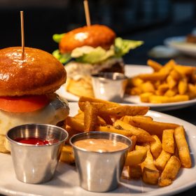 A burger sits on a tray ready to be served to customers. The burger patty is perfectly melted in white cheese and topped with a tomato between a brioche bun. Next to the burger are an assortment of seasoned fries and two dipping sauces.