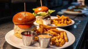 A burger sits on a tray ready to be served to customers. The burger patty is perfectly melted in white cheese and topped with a tomato between a brioche bun. Next to the burger are an assortment of seasoned fries and two dipping sauces.