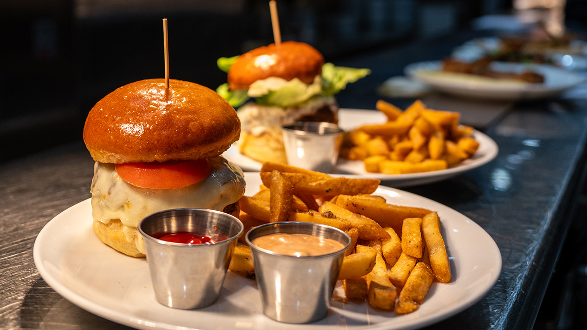 General Member Offer: 10% Off at Wolfgang Puck Bar & Grill, Disney Springs A burger sits on a tray ready to be served to customers. The burger patty is perfectly melted in white cheese and topped with a tomato between a brioche bun. Next to the burger are an assortment of seasoned fries and two dipping sauces.