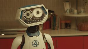 H.E.R.B.I.E., a blue and white robot from The Fantastic Four: First Steps (2025), standing in an orange and white mid-century modern kitchen.