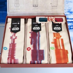 Three different pairs of socks sit in a box. Each of the socks in the box have a different lightsaber adorning them. The lightsabers all have a red overlay on them with a small Stance logo to the left, with each logo in a different color (blue, red, green). At the top left of the product image is the Star Wars logo