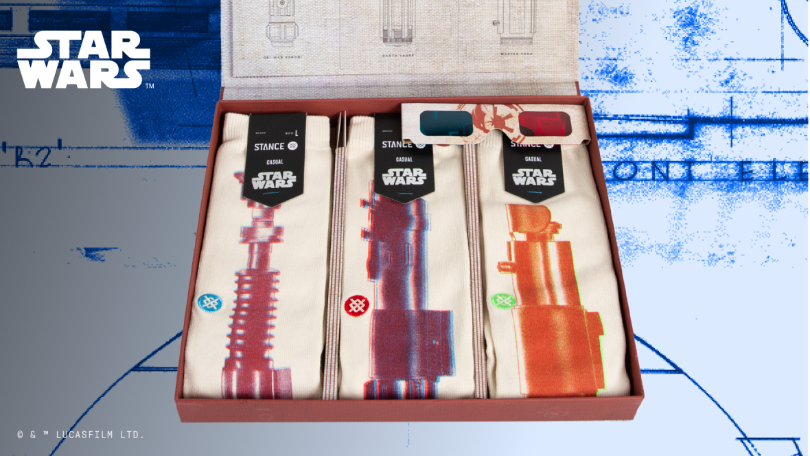 Three different pairs of socks sit in a box. Each of the socks in the box have a different lightsaber adorning them. The lightsabers all have a red overlay on them with a small Stance logo to the left, with each logo in a different color (blue, red, green). At the top left of the product image is the Star Wars logo