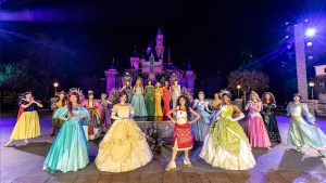 Hosts Susan Egan (left) and Ginnifer Goodwin (right) flank Disney Legend Jodi Benson, Disney Legend Anika Noni Rose, Disney Legend Paige O'Hara, Auli'i Cravalho onstage in front of Sleeping Beauty Castle at Disneyland Resort. Standing in front of them are 13 Disney Princess characters — L-R: Snow White, Raya, Ariel, Mulan, Rapunzel, Belle, Moana, Cinderella, Pocahontas, Tiana, Aurora, Merida, and Jasmine.