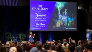 D23’s Jessica McDonald addresses an audience of D23 guests at D23 and the Walt Disney Archives’ “Disneyland: Fond Memories of the Past” Spotlight Series event in Glendale, CA. Jessica is wearing a blue dress with firework designs and stands onstage under a large projector screen of a design image of the event which features a nighttime photo of Sleeping Beauty Castle with fireworks.