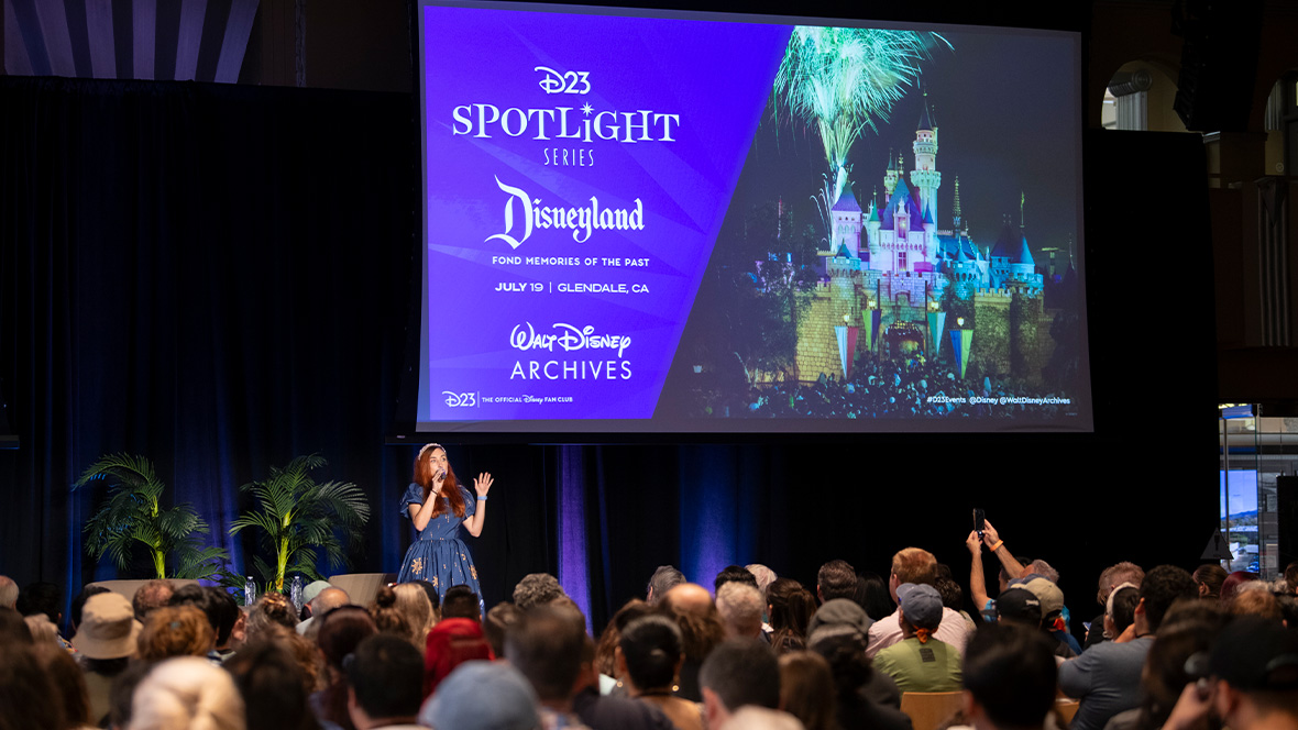 D23’s Jessica McDonald addresses an audience of D23 guests at D23 and the Walt Disney Archives’ “Disneyland: Fond Memories of the Past” Spotlight Series event in Glendale, CA. Jessica is wearing a blue dress with firework designs and stands onstage under a large projector screen of a design image of the event which features a nighttime photo of Sleeping Beauty Castle with fireworks.