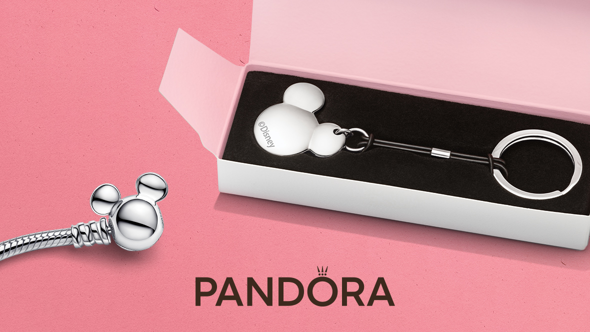 The silver Pandora clasp opener keychain sits in a white box on a light pink background. The flat clasp opener piece is shaped like Mickey Mouse with an ©Disney stamped onto it. The end of the piece is a keychain ring. The two parts are tied together by a black leather-like string. To the left of the image is the end of a silver “Disney Mickey Mouse Clasp Moments Snake Chain” bracelet, which is also shaped like Mickey Mouse. At the bottom of the image is the Pandora logo.