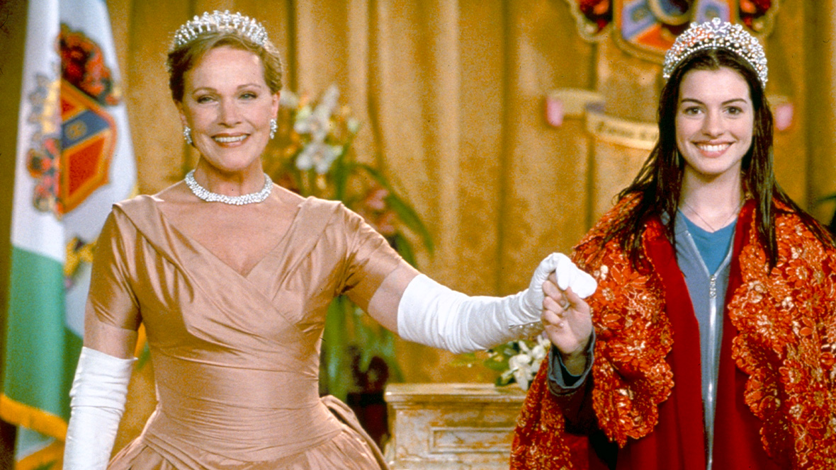 Clarisse Renaldi (Disney Legend Julie Andrews) and Mia Thermopolis (Anne Hathaway) in The Princess Diaries (2001). Clarisse is wearing a beige ballgown, white gloves, and a tiara. She is holding hands with Mia, who is wearing a red royal robe over a blue jacket and a tiara.