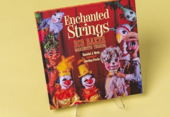 A hardcover book sits against a light-yellow background. The text on the cover of the book reads “Enchanted Strings, Bob Baker Marionette Theater, Randal J. Metz, Forward by Jordan Peele.” Surrounding this text are assorted puppets from Bob Baker Marionette Theater—including multiple clowns, a bird, hybrid-plant puppets, and more. The text and puppets are placed in front of a red stage curtain. 