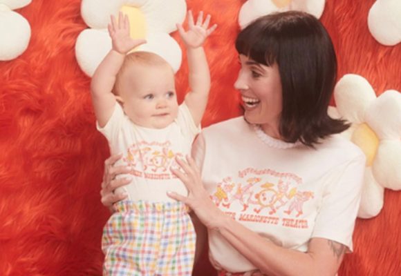 A smiling woman is holding a baby, and they’re standing in front of a fuzzy orange background adorned with plushie flowers. Both are wearing matching Bob Baker Marionette Theater shirts featuring line art of a puppet marching band. The baby holds their hands up in the air.