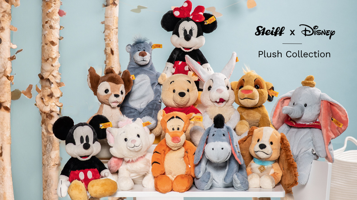 Group of Steiff plush toys from the Disney Plush Collection, including Mickey Mouse, Minnie Mouse, Winnie the Pooh, Tigger, Eeyore, Dumbo, Simba, Lady, Baloo, Marie, Bambi, and Thumper, posed in front of birch trees and a soft blue background.