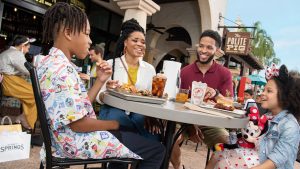 A family sits around an outdoor table, enjoying a meal, at The Polite Pig at Disney Springs at Walt Disney World Resort. From left, we see a boy in a Disney print short-sleeve shirt, a mom in a white jacket with yellow shirt beneath, a dad in a maroon pullover, and a little girl in a white Disney-theme dress worn beneath a denim jacket. The girl is holding a plush Minnie Mouse. The table is set with trays holding food, and a roll of paper towels in a dispenser.