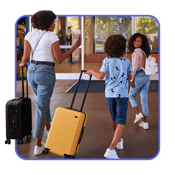 Hotels Woman and two children walk into a building with rolling suitcases, one child wearing a Mickey Mouse shirt and another smiling back toward the camera.