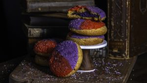 A stack of three Gideon&rsquo;s specialty D23 Coconut Ube Sugar Cookies sit elevated on a wooden display. The sprinkle and colors of the top of the cookie are split in half. One side is purple, while the other is red.