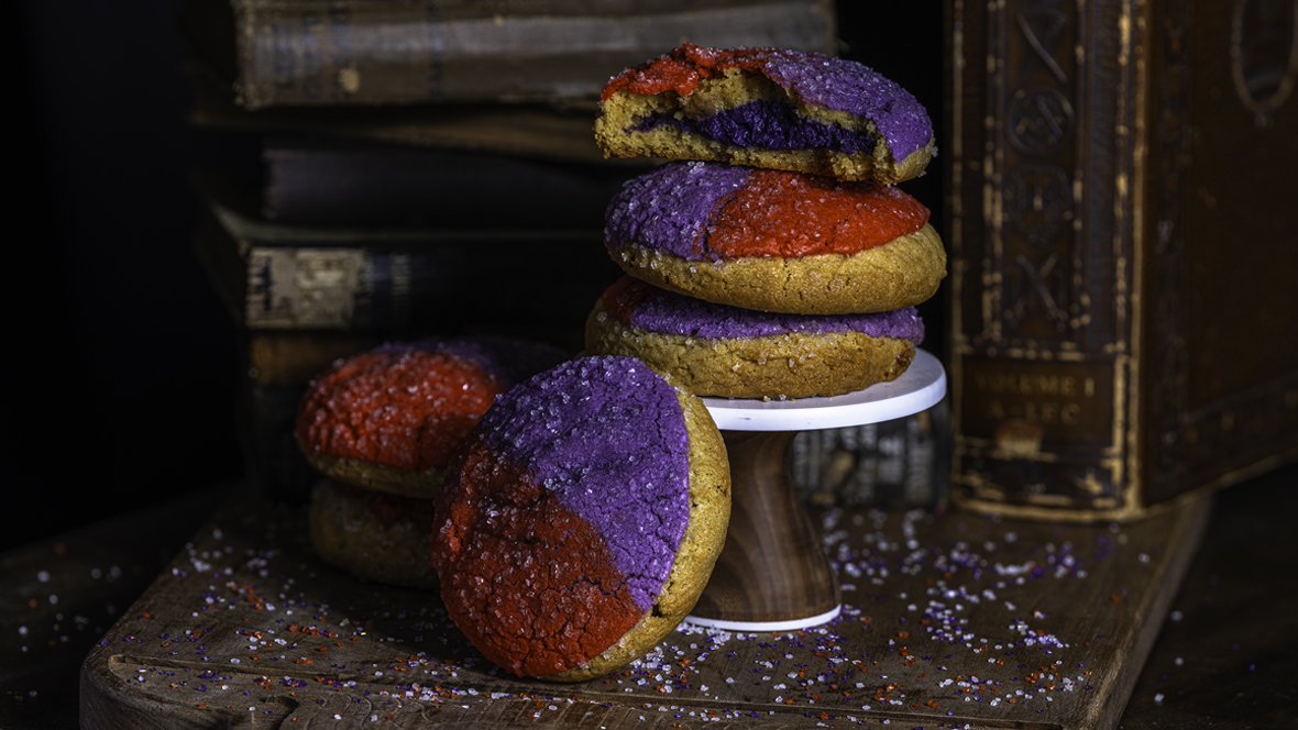 A stack of three Gideon’s specialty D23 Coconut Ube Sugar Cookies sit elevated on a wooden display. The sprinkle and colors of the top of the cookie are split in half. One side is purple, while the other is red.