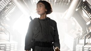 Wendy (Sydney Chandler) from FX’s Alien: Earth is seen wearing a grey jumpsuit and standing in front of a brightly lit hallway. She is frightened, looking at something in the distance.