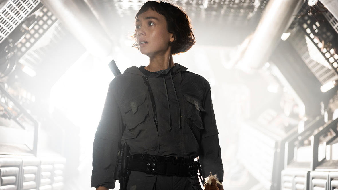 Wendy (Sydney Chandler) from FX’s Alien: Earth is seen wearing a grey jumpsuit and standing in front of a brightly lit hallway. She is frightened, looking at something in the distance.