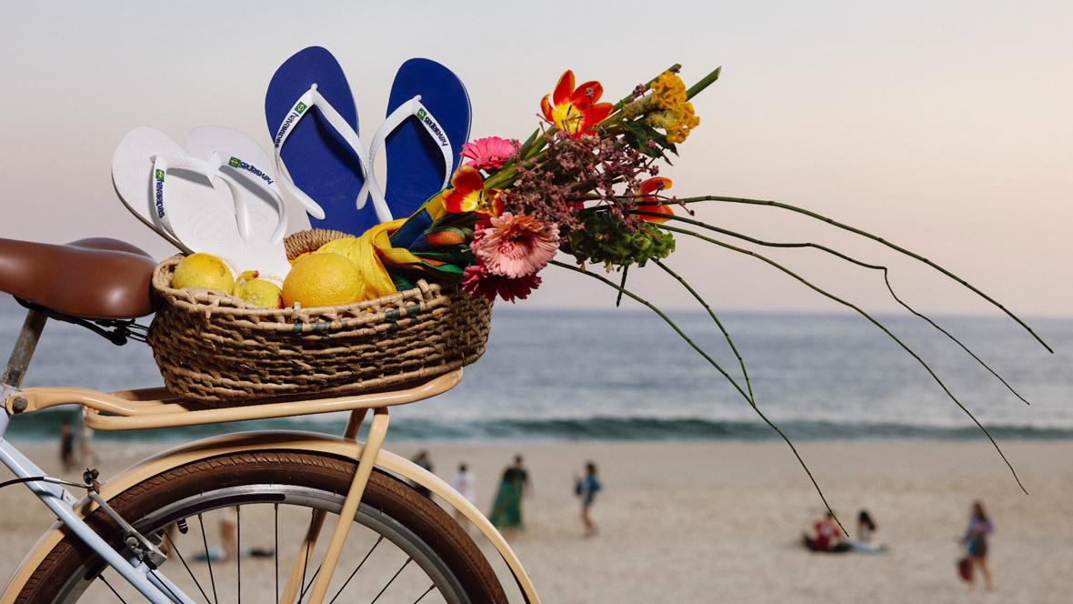  Close up shot of two pairs of flip flops accompanied by florals and fruit in a bicycle basket sitting in front of a beach.