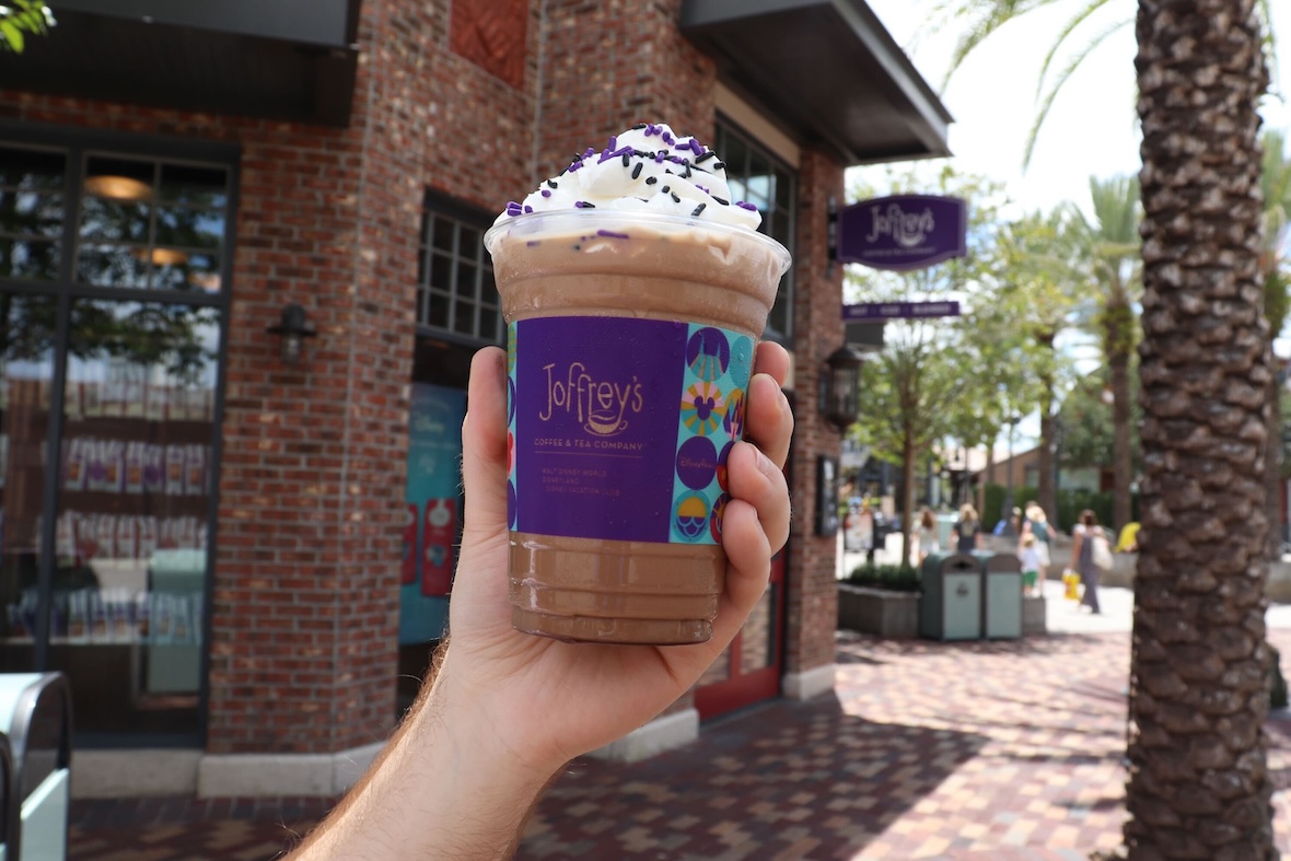 Hand holding a Joffrey’s Coffee & Tea Company frozen drink topped with whipped cream and purple sprinkles in front of the Joffrey’s storefront at Disney Springs.