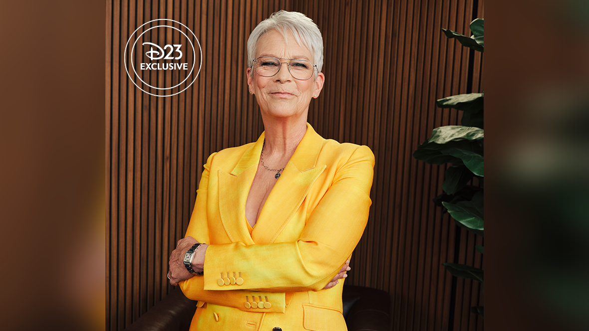 Jamie Lee Curtis, the star of Freaky Friday, poses for a portrait in front of an unadorned wall made up of wooden slats. Curtis stands with her arms crossed at her waist, wearing  a bright canary yellow suit jacket and metal rimmed eyeglasses. Beneath the jacket, barely visible, is a yellow V-neck shirt. Her hair, cut fairly short, is gray and nearly white. She has a watch on her left wrist and a thin necklace with a simple pendant. She is smiling broadly with her mouth closed. The arms of a leather chair are visible behind her.