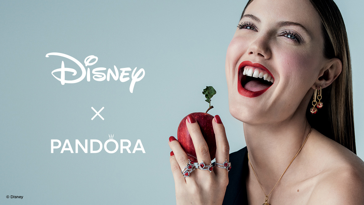 Smiling woman holding a red apple, wearing red and gold Disney x Pandora jewelry inspired by Disney's Snow White, with Disney and Pandora logo on a light blue background.