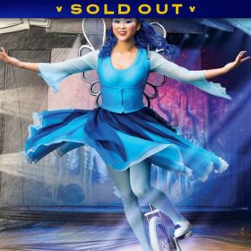 A promotional image for Drawn to Life presented by Cirque du Soleil and Disney at Disney Springs in Orlando, Florida. The left side of the image features a hand-drawn theater, with rows of seats sketched by pencil with monochrome shading. The right side of the image transitions from the hand-drawn style into a full color photo of a theater stage. The stage is framed by large pages from a sketchbook, and as the image moves from left to right, the sketches move from a hand-drawn style to being in color. In the foreground of the image is a Drawn to Life performer and character, representing the Blue Fairy from Pinocchio during a unicycle act. The female-presenting character is in a flowy dress in shades of blue and wearing a dark blue wig and silver wings. While on the unicycle, the character evokes action and grace. The D23 logo is in the bottom right corner in white. A banner at the top reads SOLD OUT.