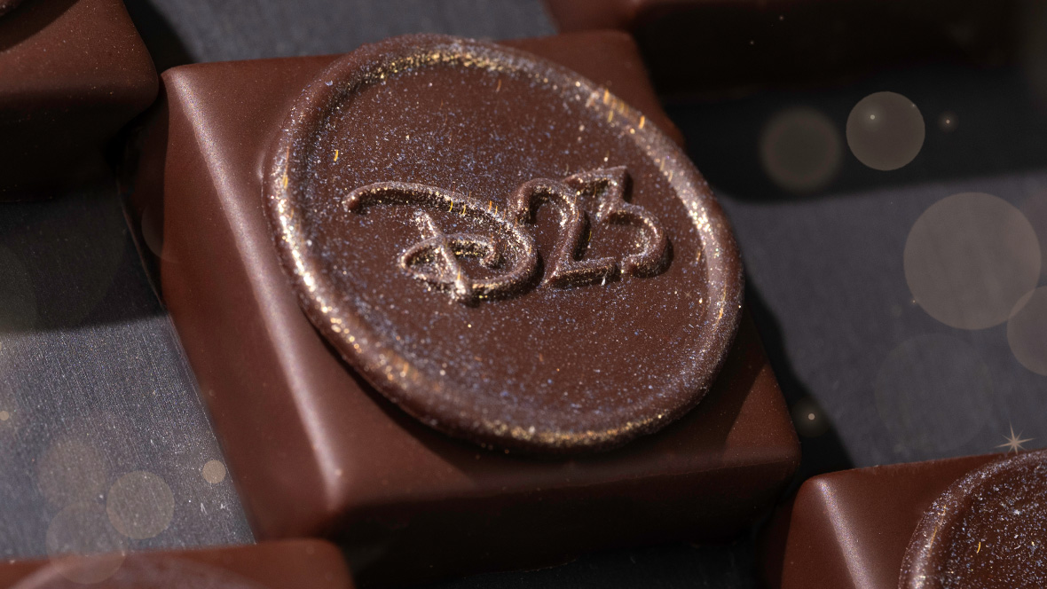 Specialty D23 Chocolate Square at The Ganachery, Disney Springs A close-up photo of a delectable chocolate square sits on a gray serving tray. The chocolate treat is topped with an edible circle stamped with the D23 logo. The edible circle is dusted with a golden shimmer to make it shine in the light.