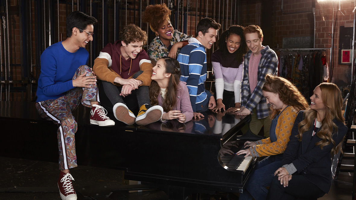 The cast of the High School Musical: The Musical: The Series (2019-2023) sits around a piano. (From left to right) Carlos (Frankie A. Rodriguez), Ricky (Joshua Basset), Nini (Olivia Rodrigo), Kourtney (Dara Reneé), EJ (Matt Cornett), Gina (Sofia Wylie), Seb (Joe Serafini), Ashlyn (Julia Lester), and Miss Jenn (Kate Reinders).
