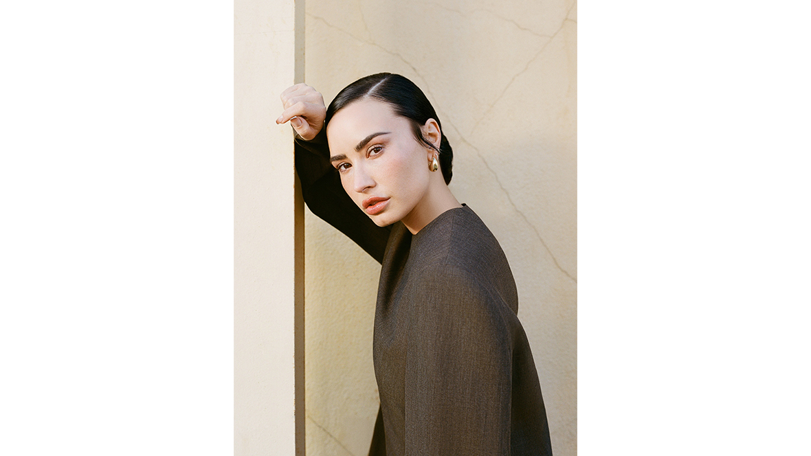 Demi Lovato, photographed by Jane Dylan Cody, poses against a wall. Her brownish-black hair is slicked back and she is wearing a dun-colored top and gold earrings.