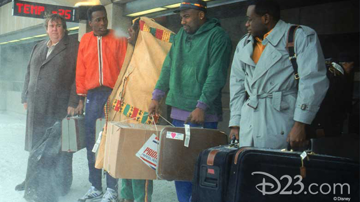 In a still from Disney’s Cool Runnings, beloved comedian John Candy stands with four actors playing members of the Jamaica national bobsled team in a snowy scene. The bobsled team looks miserable and cold. 
