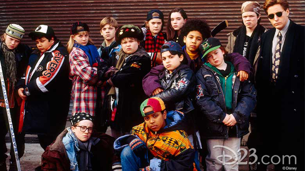 Emilio Estevez, who stars as Gordon Bombay in Disney’s The Mighty Ducks, stands in sunglasses and office attire with the cast of relative newcomers try to look tough. 