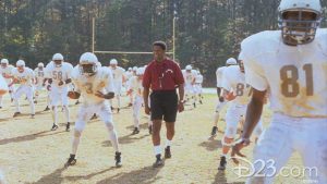 Denzel Washington, playing coach Herman Boone, in Remember the Titans stands smiling in a red polo, black shorts with a whistle around his neck as football players dressed in white uniforms practice.