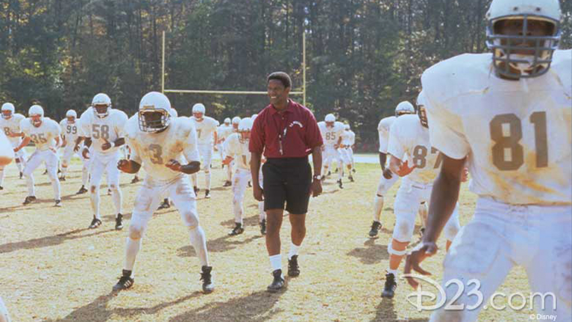 Denzel Washington, playing coach Herman Boone, in Remember the Titans stands smiling in a red polo, black shorts with a whistle around his neck as football players dressed in white uniforms practice.