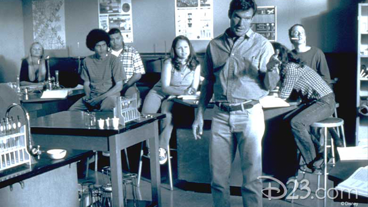 In a still from Disney’s The Rookie, Dennis Quaid as Jim Morris, stands in the middle of his high school classroom mid-instruction as several students look on.