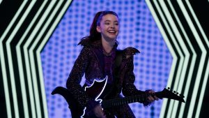 Vee (played by Kenzi Richardson) from Vampirina: Teenage Vampire stands onstage wearing a purple dress and playing a black, bat-shaped guitar.