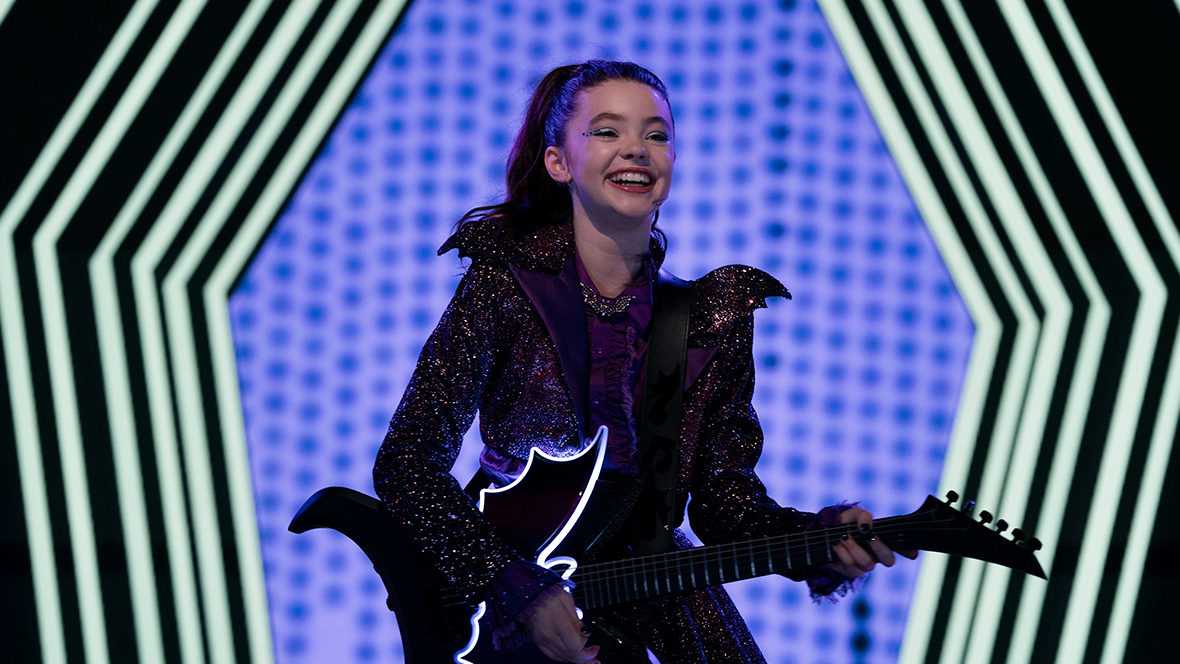 Vee (played by Kenzi Richardson) from Vampirina: Teenage Vampire stands onstage wearing a purple dress and playing a black, bat-shaped guitar.