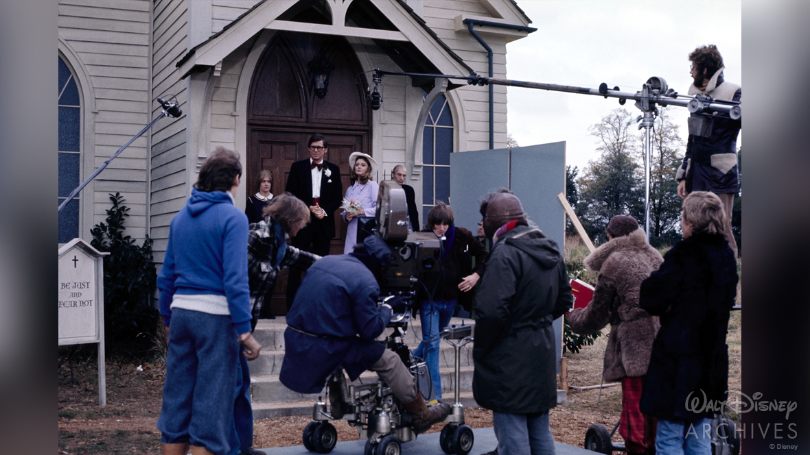Behind the scenes. On the steps of the church are Patricia Quinn, Barry Bostwick, Susan Sarandon, and Richard O’Brien. Bostwick and Sarandon are in formal clothing, Quinn and O’Brien look like members of the church. The film production crew is in the foreground with cameras and boom microphones.