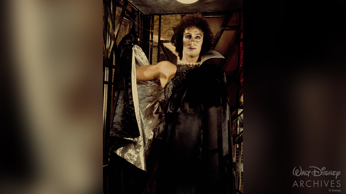 Actor Tim Curry as Dr. Frank-N-Furter in The Rocky Horror Picture Show, He has curly black hair is wearing glamorous makeup and a black cape, and is standing in an elevator.