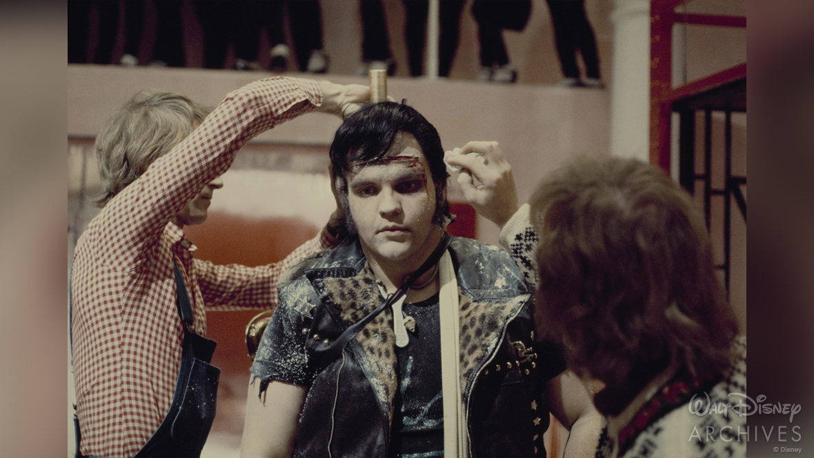 Meat Loaf (Eddie) getting his makeup retouched on the set of The Rocky Horror Picture Show. He sports a black pompadour hairstyle, and two crew members surround him, applying makeup and styling his hair.