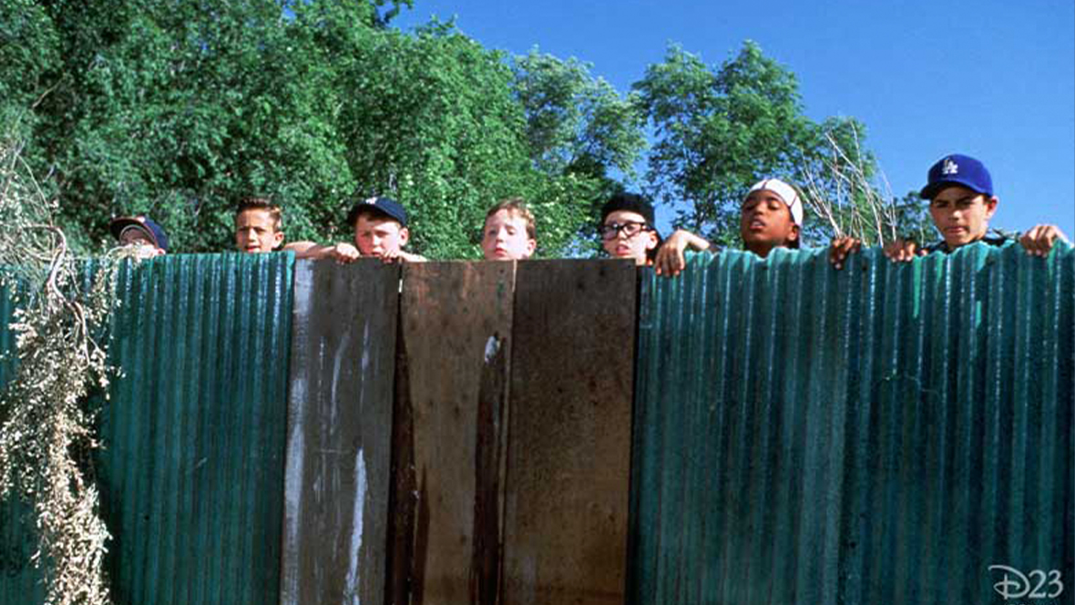 In a still from Disney’s The Sandlot, seven boys with just their heads and hands visible, peer over an alternating medal and wood wall with a blue sky and green trees behind them. The boy at far right wears a blue LA Dodgers baseball cap. 